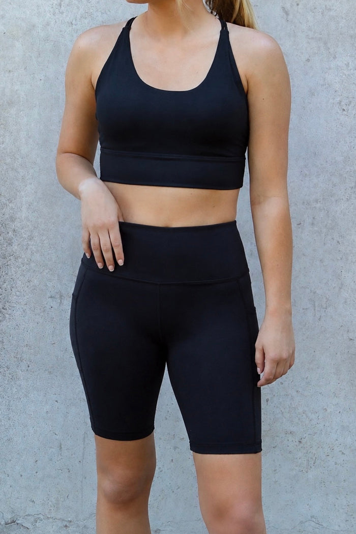 A woman modeling a matching black activewear set consisting of a scoop-neck sports bra and high-waisted biker shorts, standing against a textured gray concrete wall.