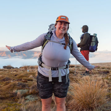 A woman hiking on a mountain ridge with her arms outstretched, wearing a light gray long-sleeve top, dark shorts, and an orange cap, with another hiker in the background under a clear blue sky.