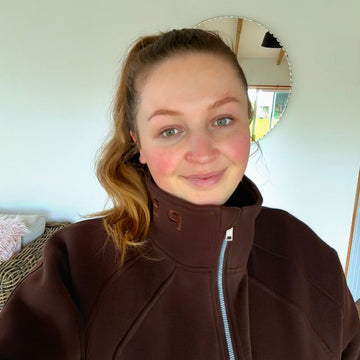A woman wearing a dark brown zip-up fleece jacket with a high collar, photographed in a casual indoor setting with a white wall and a round mirror behind her.