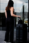A woman stands with her back to the camera, wearing a black sleeveless top and black wide-leg pants with a white side stripe. She is holding the handle of a black hard-shell suitcase next to a black handbag in front of a modern glass building.