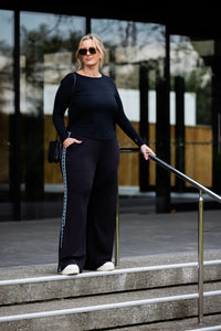 A woman stands on outdoor concrete steps in front of a modern glass building, wearing a black long-sleeve top and matching black wide-leg pants with a light blue stripe detail down the side, paired with white sneakers and a black shoulder bag.