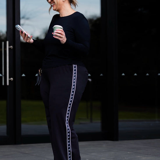 A woman stands outdoors in front of large black-framed glass doors, wearing a black long-sleeve top and black wide-leg pants with a white decorative stripe down the side. She holds a white coffee cup and looks at her phone.