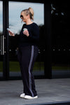 A woman stands outdoors in front of large black-framed glass doors, wearing a black long-sleeve top and black wide-leg pants with a white decorative stripe down the side. She holds a white coffee cup and looks at her phone.