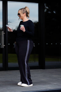 A woman stands outdoors in front of large black-framed glass doors, wearing a black long-sleeve top and black wide-leg pants with a white decorative stripe down the side. She holds a white coffee cup and looks at her phone.