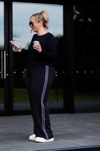 A woman stands outdoors in front of large black-framed glass doors, wearing a black long-sleeve top and black wide-leg pants with a white decorative stripe down the side. She holds a white coffee cup and looks at her phone.
