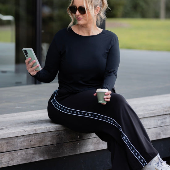A woman sits on a wooden bench outdoors, wearing a black long-sleeve top and matching black wide-leg pants with a light blue geometric stripe detail. She is holding a phone and a coffee cup.