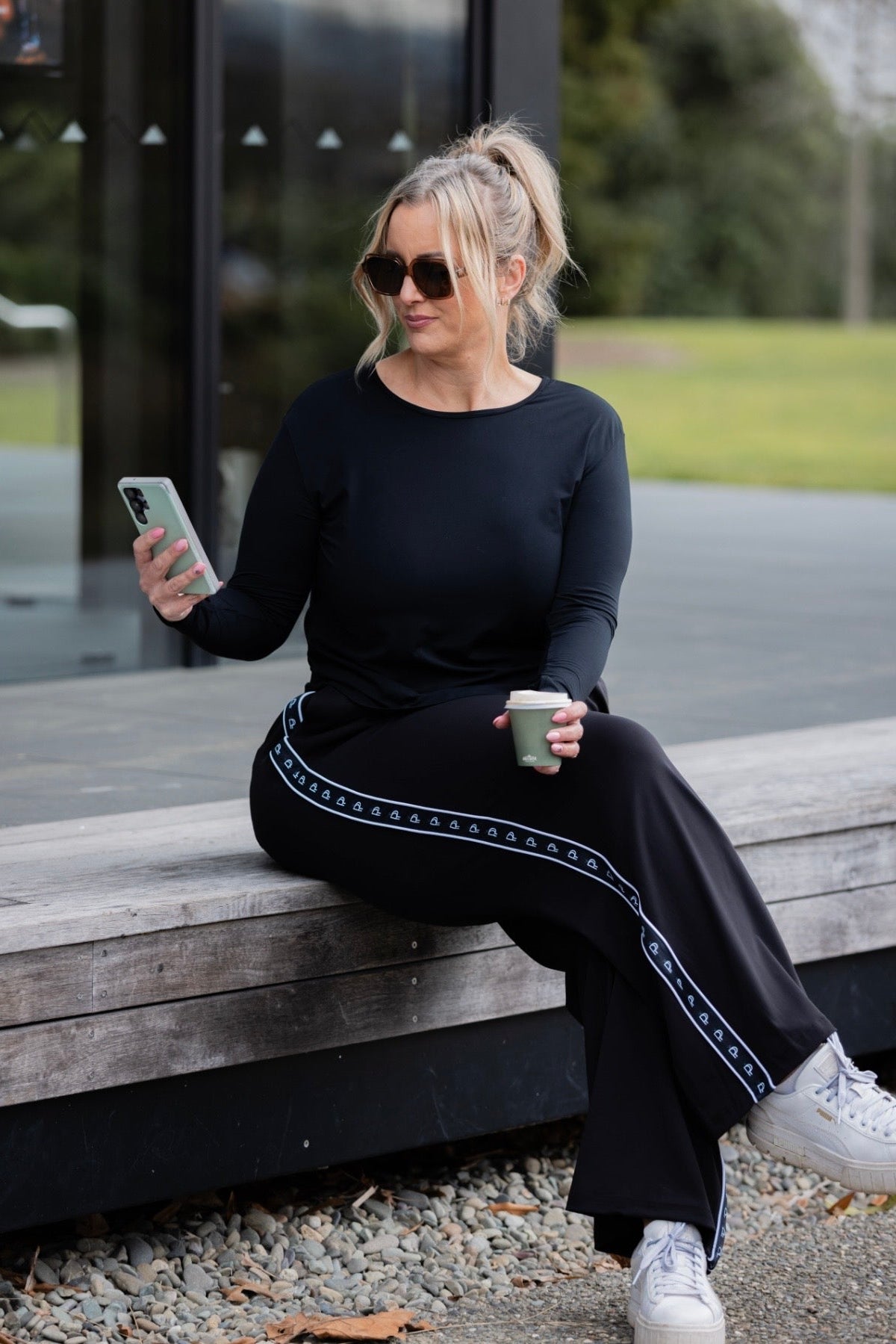 A woman sits on a wooden bench outdoors, wearing a black long-sleeve top and matching black wide-leg pants with a light blue geometric stripe detail. She is holding a phone and a coffee cup.
