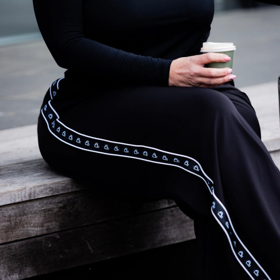 A person seated on a wooden bench wearing a black long-sleeve top and black wide-leg pants with a white decorative stripe running down the side. The person is holding a takeaway coffee cup.