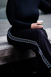 A person seated on a wooden bench wearing a black long-sleeve top and black wide-leg pants with a white decorative stripe running down the side. The person is holding a takeaway coffee cup.