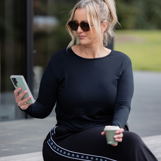 A woman wearing a black long-sleeve top and black wide-leg trousers with a white geometric stripe detail sits on a wooden bench outdoors, holding a phone and a coffee cup.