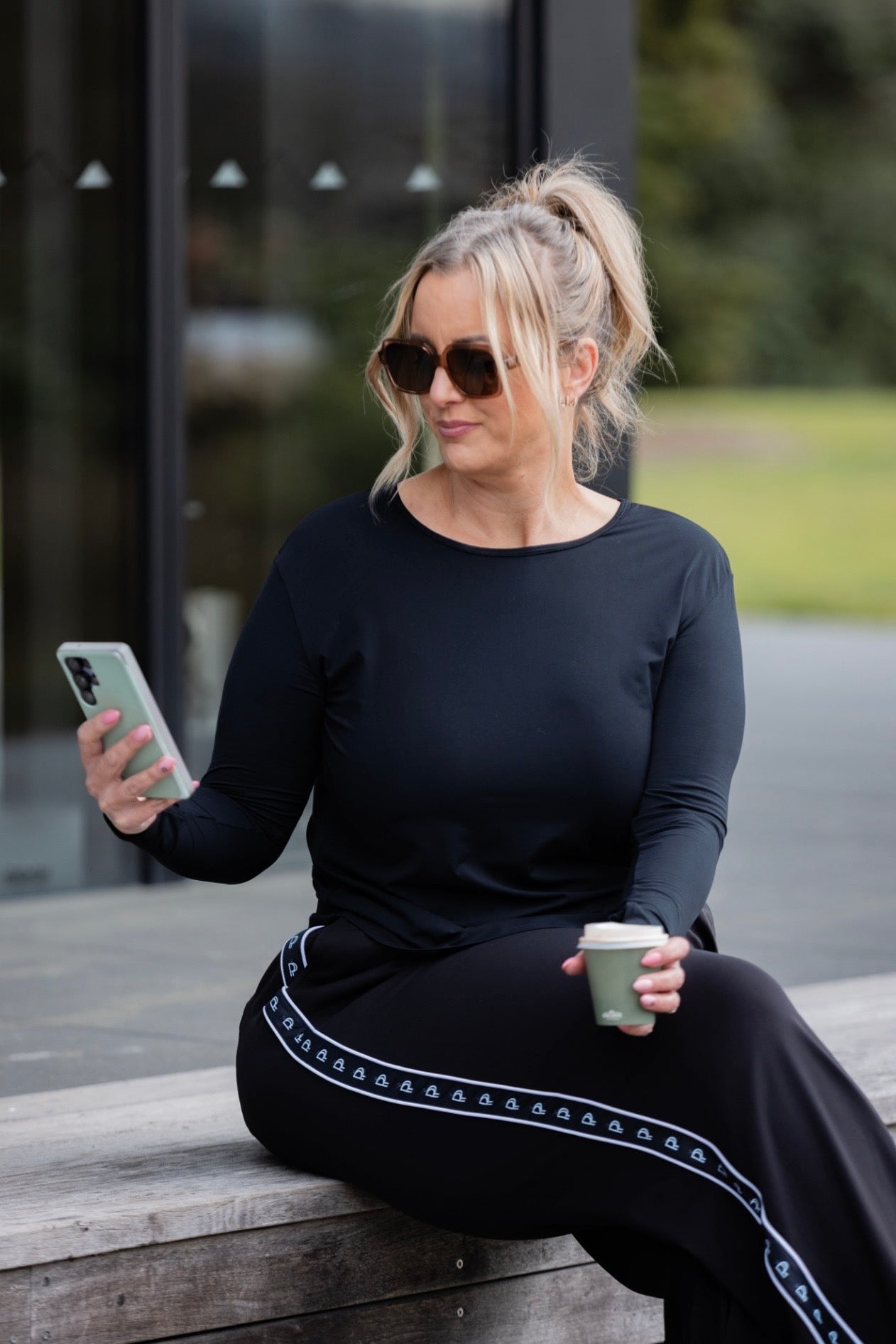 A woman wearing a black long-sleeve top and black wide-leg trousers with a white geometric stripe detail sits on a wooden bench outdoors, holding a phone and a coffee cup.