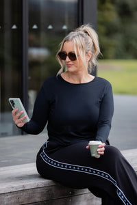 A woman wearing a black long-sleeve top and black wide-leg trousers with a white geometric stripe detail sits on a wooden bench outdoors, holding a phone and a coffee cup.