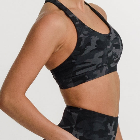 A woman modeling a matching black and gray camouflage activewear set, featuring a sports bra and high-waisted biker shorts, against a plain white studio background.