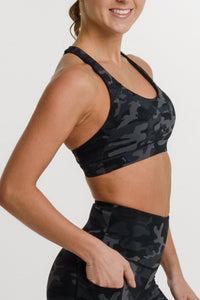 A woman modeling a matching black and gray camouflage activewear set, featuring a sports bra and high-waisted biker shorts, against a plain white studio background.