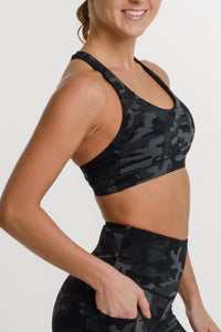 A woman modeling a matching black and gray camouflage activewear set, featuring a sports bra and high-waisted biker shorts, against a plain white studio background.