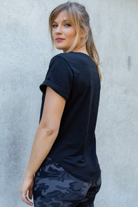 Woman wearing a black t-shirt and camouflage pants against a gray wall