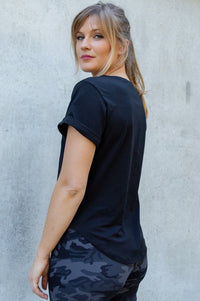 Woman wearing a black t-shirt and camouflage pants against a gray wall