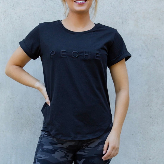Woman wearing a black t-shirt with 'Peche' branding and camouflage shorts against a light gray wall.