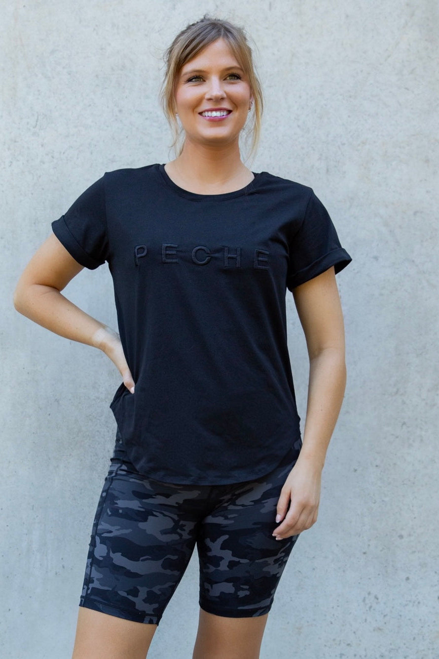 Woman wearing a black t-shirt with 'Peche' branding and camouflage shorts against a light gray wall.