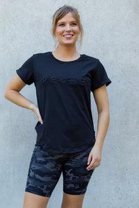 Woman wearing a black t-shirt with 'Peche' branding and camouflage shorts against a light gray wall.