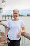 Woman in a white t-shirt standing on a wooden deck by a waterfront.
