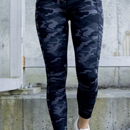 Person wearing dark grey/ black camouflage leggings with white sneakers on a concrete surface.
