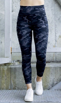 Person wearing dark grey/ black camouflage leggings with white sneakers on a concrete surface.