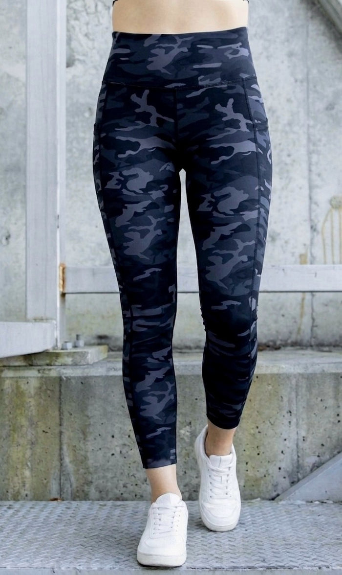 Person wearing dark grey/ black camouflage leggings with white sneakers on a concrete surface.