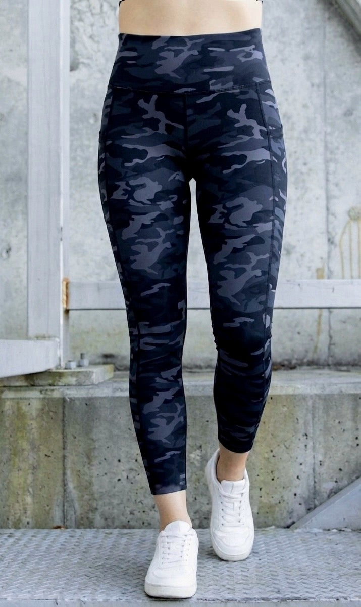 Person wearing dark grey/ black camouflage leggings with white sneakers on a concrete surface.