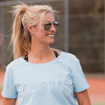 A woman on a tennis court wearing a light blue short-sleeve t-shirt with the brand name 'PECHE' printed across the chest in a matching tone.
