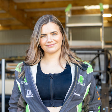 A woman with long brown hair stands in a workshop wearing a black and green racing jacket over a black top. The jacket features green and white graphics.