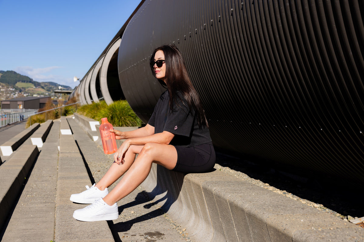A woman wearing a black t-shirt and black shorts sits on concrete steps next to a large black corrugated metal structure, holding an orange water bottle.