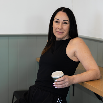 A woman with long dark hair sits in a wooden booth, wearing a sleeveless black top and black sweatpants with a white side stripe. She is holding a white paper coffee cup.