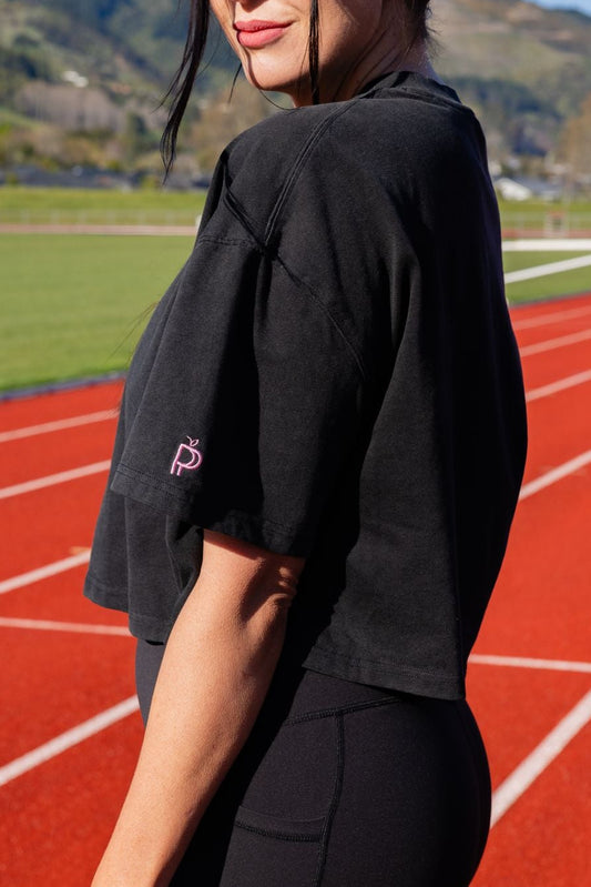 A woman wearing a black cropped t-shirt and black leggings stands on a red running track in an outdoor stadium with green fields and hills in the background.