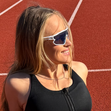 A woman on a red running track wearing white-framed shield sunglasses with dark lenses, a black athletic top, and a silver chain necklace.