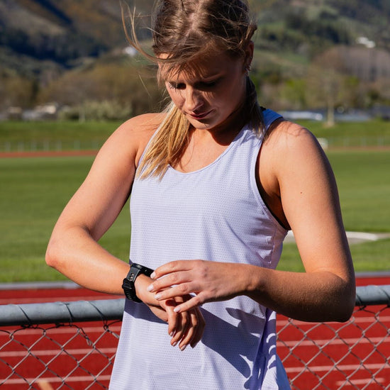 A woman on an outdoor running track checks her smartwatch. She is wearing a light lavender sleeveless athletic tank top and dark gray camouflage running shorts, with a chain-link fence and green grassy field in the background, and mountains under a blue sky further back.
