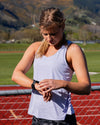 A woman on an outdoor running track checks her smartwatch. She is wearing a light lavender sleeveless athletic tank top and dark gray camouflage running shorts, with a chain-link fence and green grassy field in the background, and mountains under a blue sky further back.