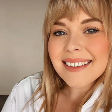 A close-up portrait of a smiling blonde woman wearing a white satin zip-up robe against a plain beige wall.
