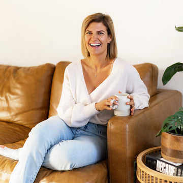 Woman sitting on a brown leather couch holding a white mug in a cozy living room.