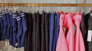 A clothing rack displaying multiple activewear garments in different colors, including navy blue, black, and bright pink, hanging on a gold metal rod.