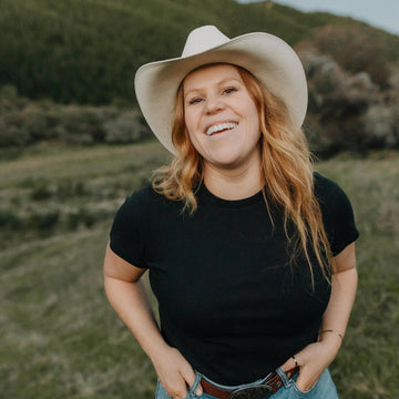 A smiling woman outdoors wearing a white cowboy hat, black short-sleeve t-shirt, and blue jeans, standing in a grassy field with hills in the background.