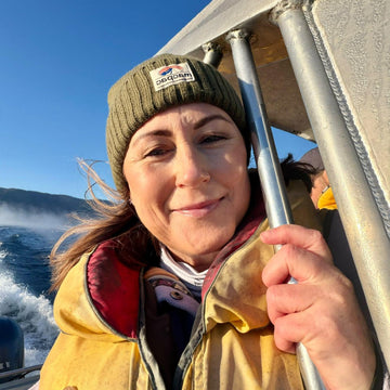 A woman on a boat wearing a green knit beanie and a yellow life jacket, with blue ocean and sky in the background.